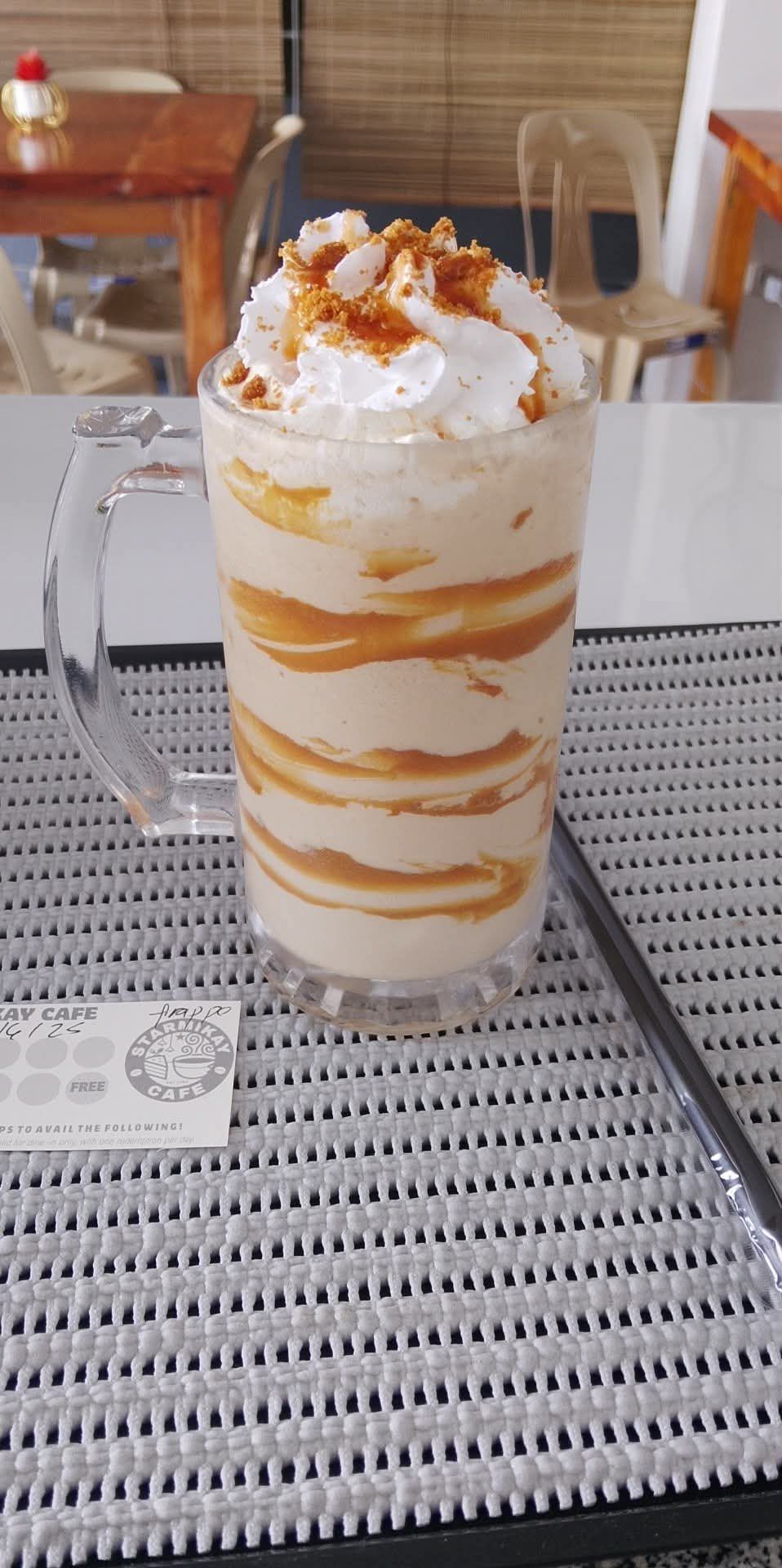 A tall glass mug filled with a creamy, caramel-swirled blended drink, topped with whipped cream on a patterned mat.
