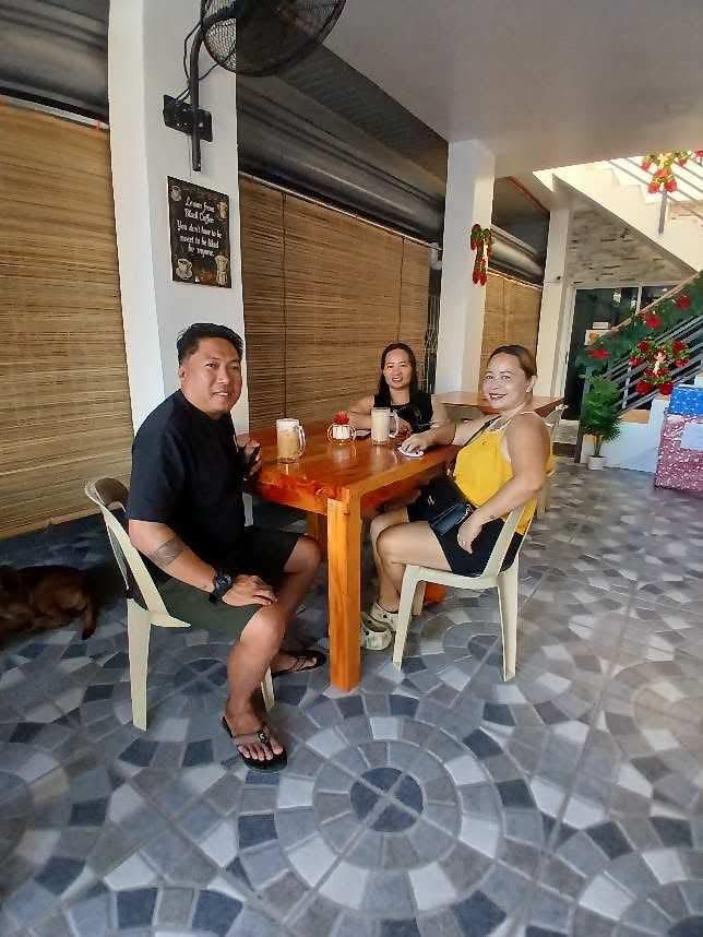 Three people sit at a wooden table in an outdoor dining area with a tiled floor, smiling at the camera.