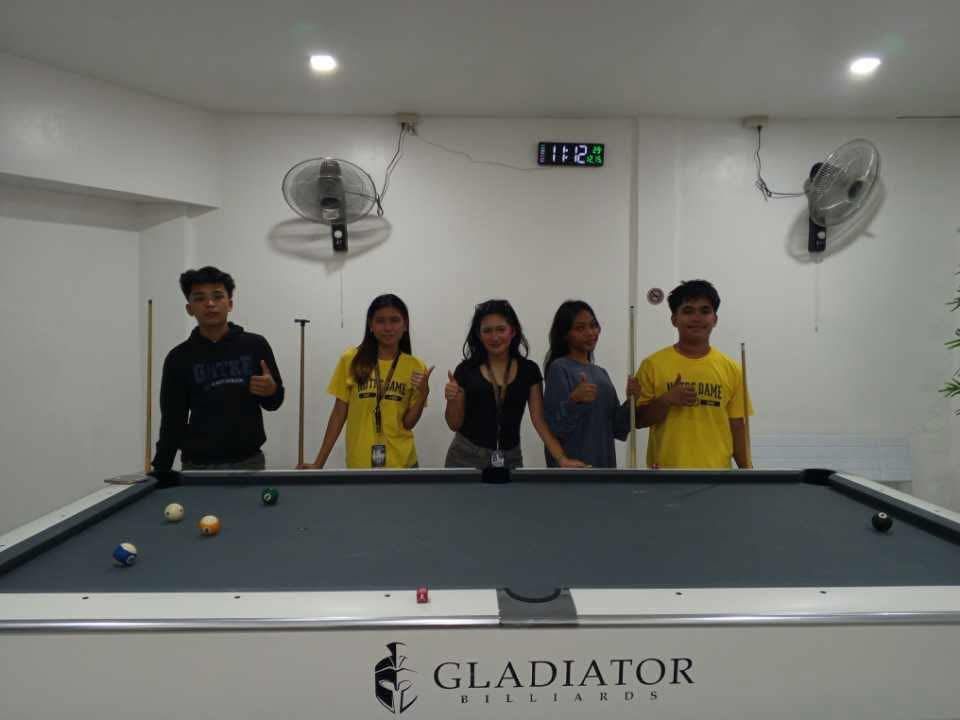 Five people stand behind a pool table in a white room, smiling and posing for a photo.
