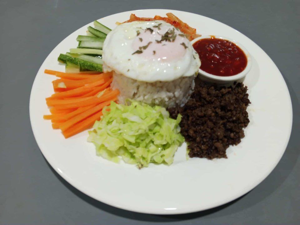 A white plate served with rice topped with a fried egg, surrounded by julienned carrots, cucumber, cabbage, beef, and kimchi.