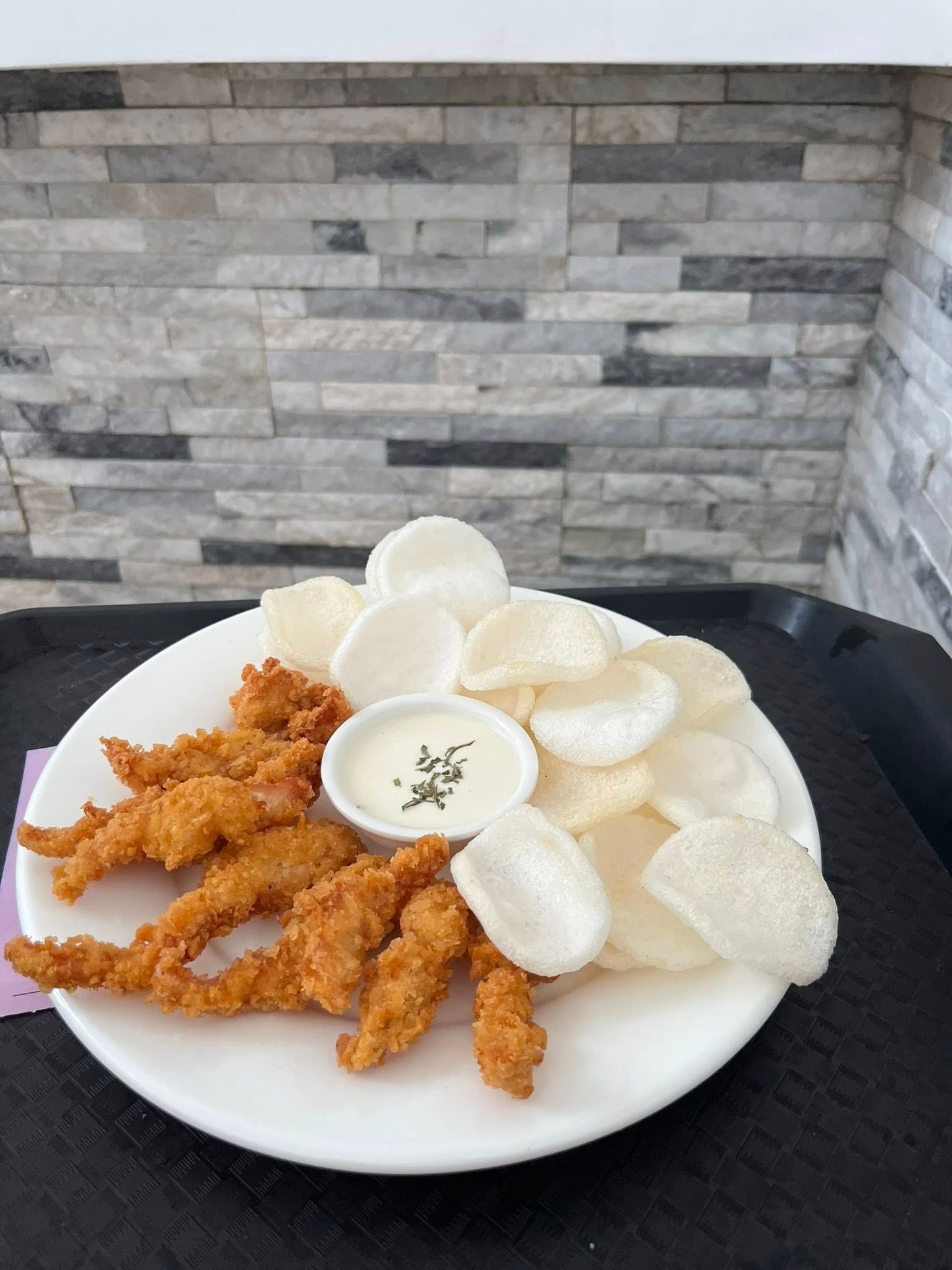 A plate of crispy breaded chicken tenders served with white prawn crackers and a small bowl of creamy dipping sauce.