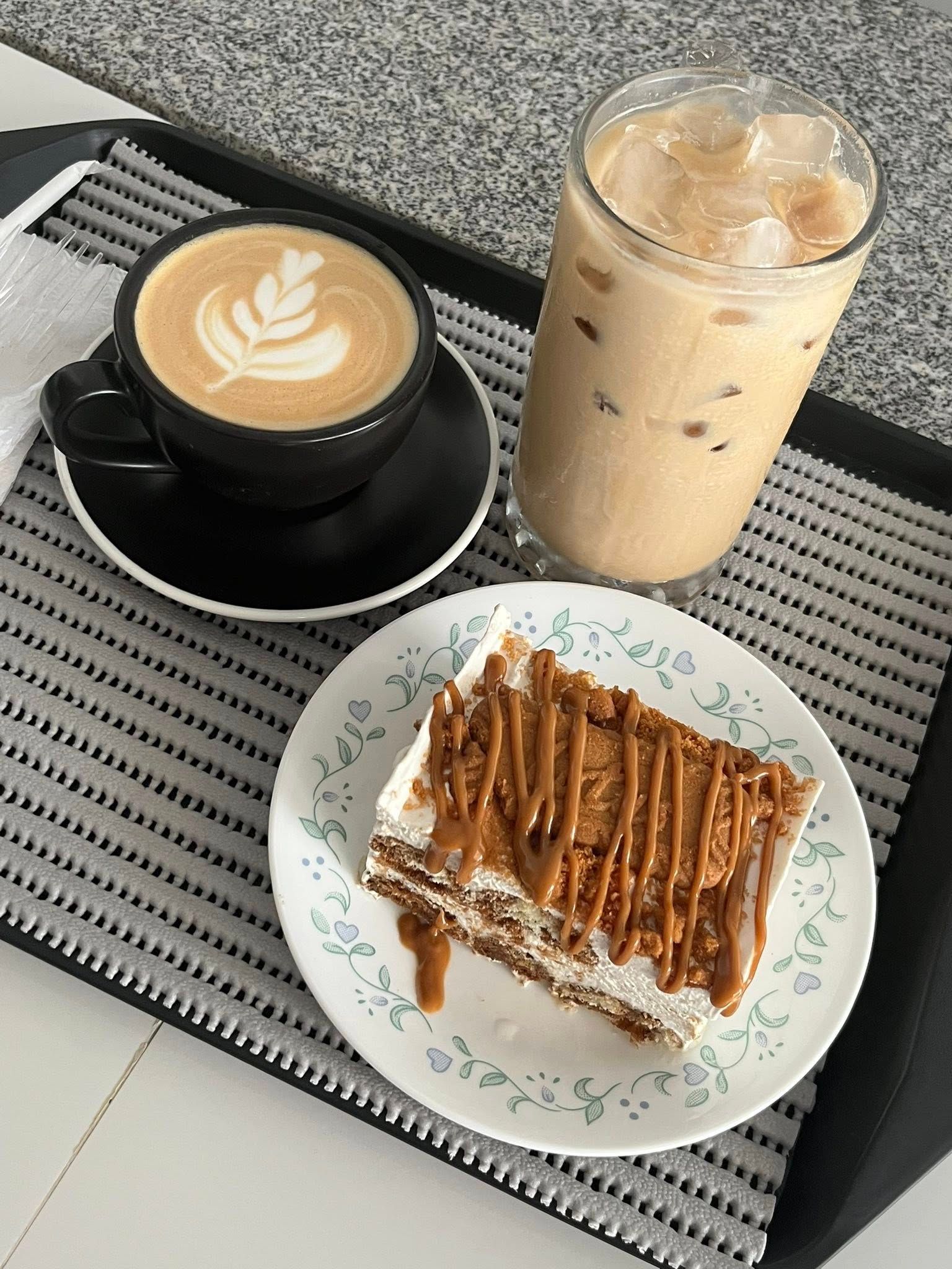 A tray holds a hot latte with latte art, an iced coffee in a tall glass, and a square slice of cake with caramel drizzle.