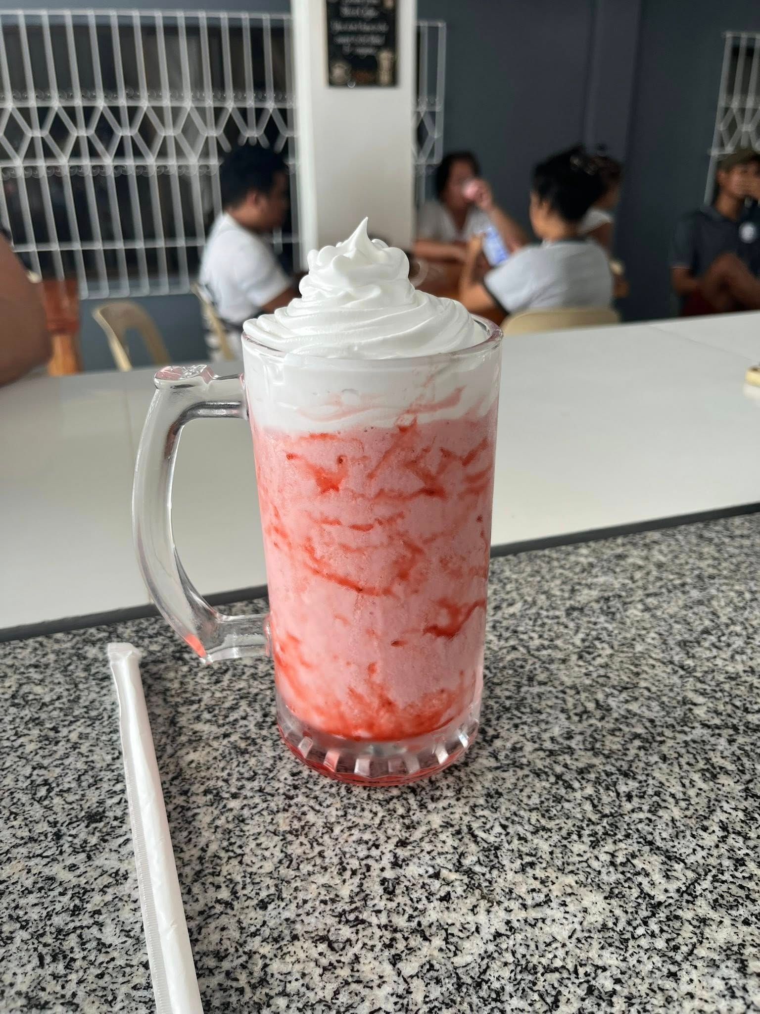 A tall glass mug filled with a pink strawberry milkshake topped with whipped cream, sitting on a speckled cafe table.