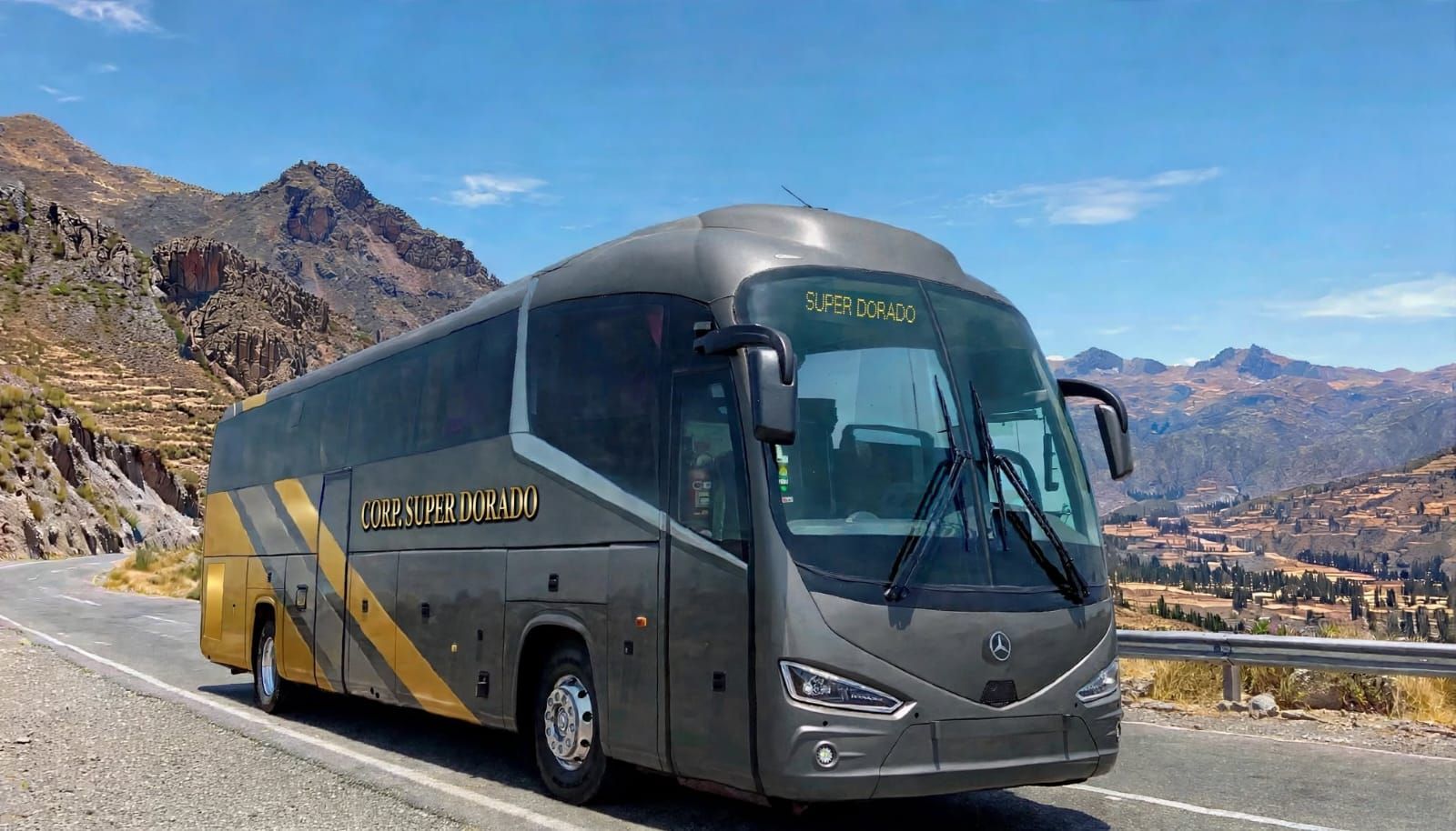 Autobús de pasajeros blanco y azul circulando por una carretera de montaña con colinas rocosas al fondo.