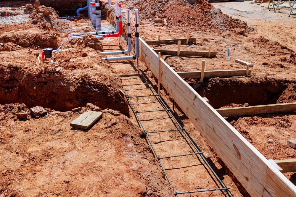 Construction site with trench forms, rebar, and plumbing pipes on muddy ground