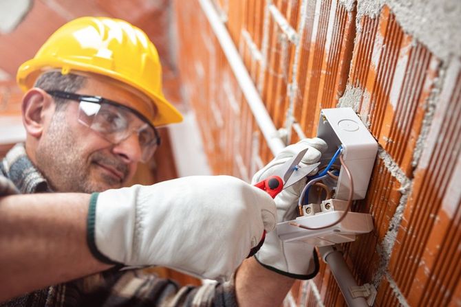 Electrician installing an outlet in a wall, wearing a yellow hard hat and safety glasses