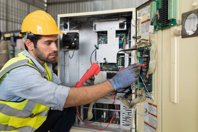 Technician in hard hat and safety vest repairing an open electrical control panel with tools.