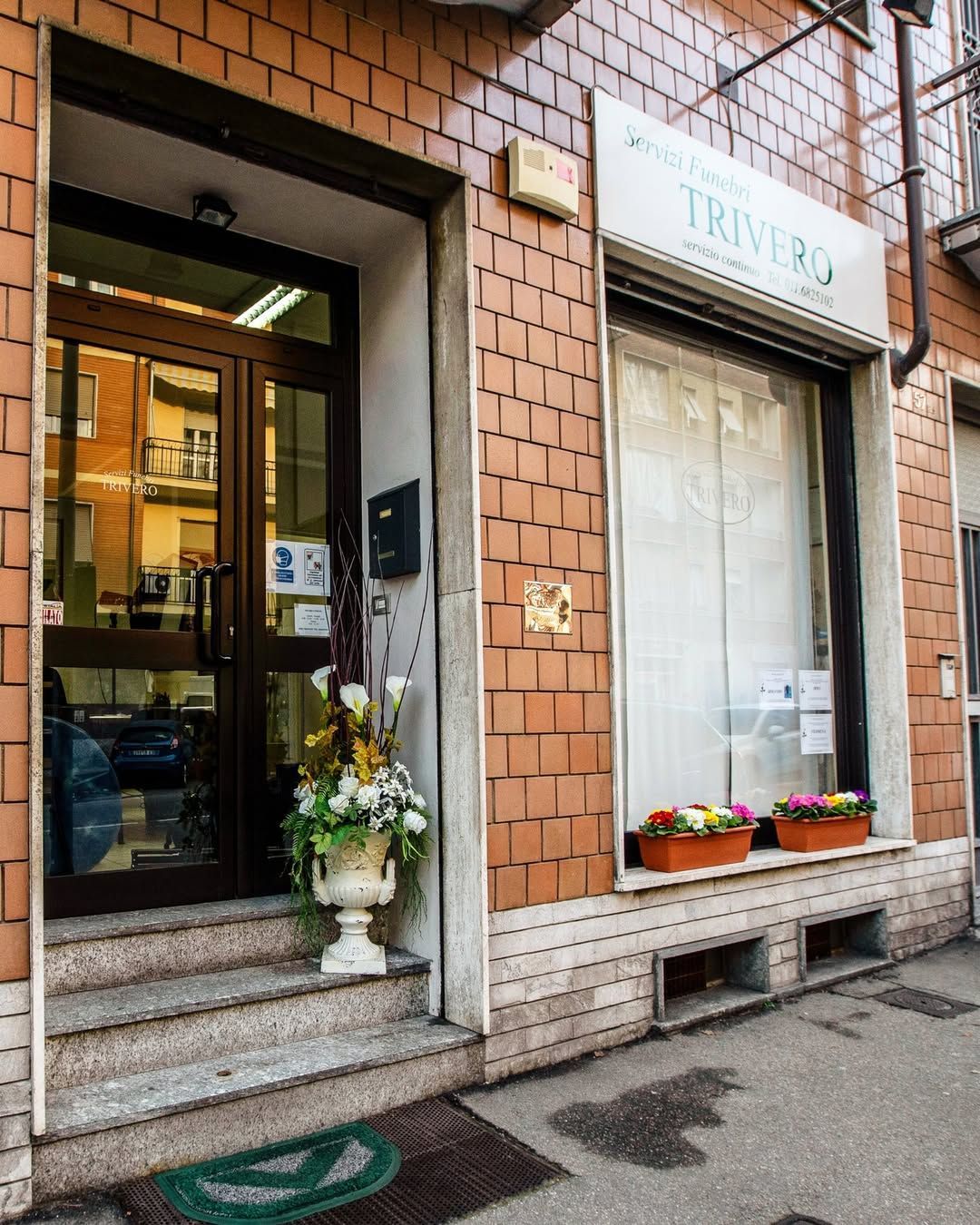 Vetrina con ingresso aperto, tenda da sole bianca e fioriere su un marciapiede in mattoni.