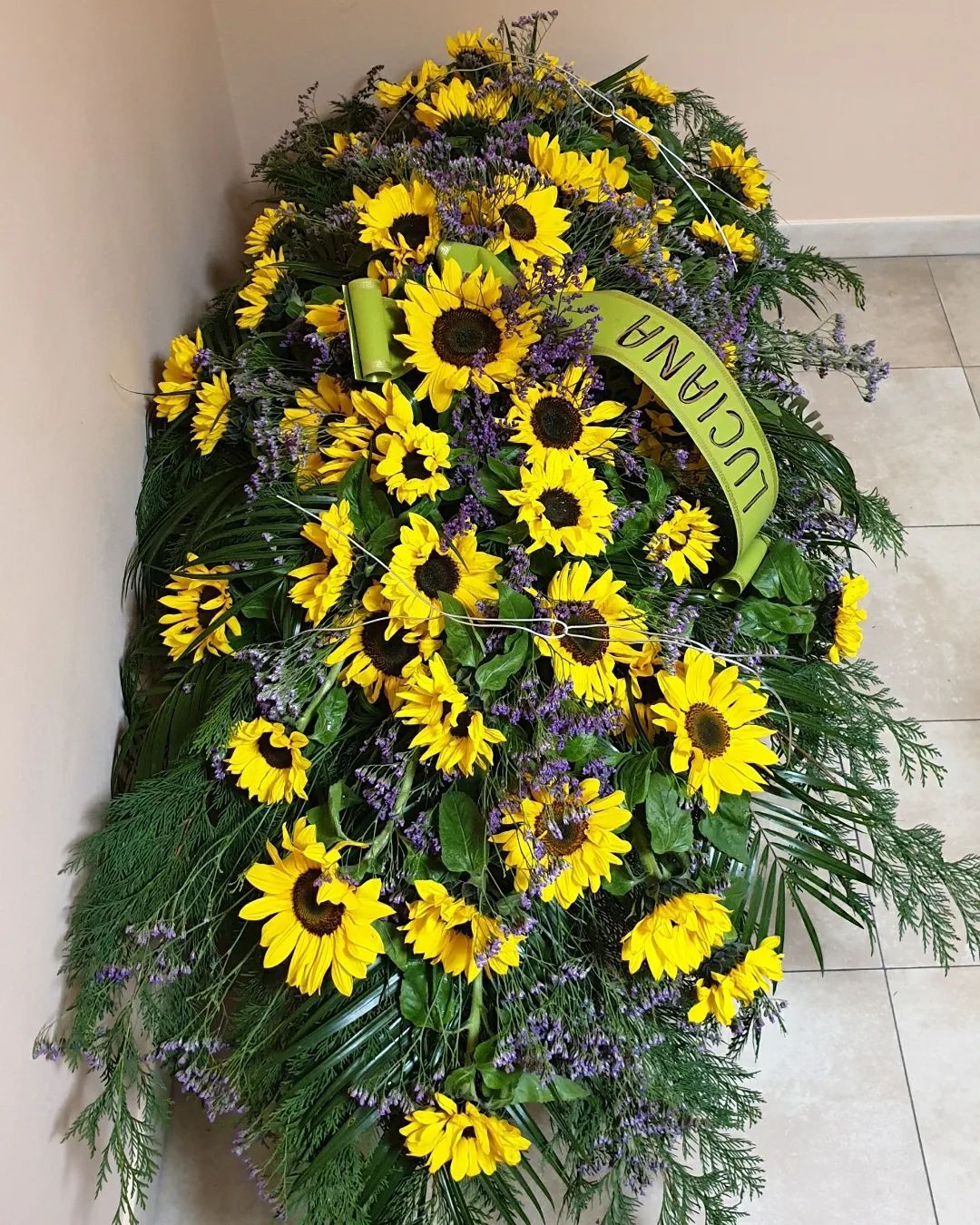 Composizione floreale funebre gialla e viola con nastro verde, esposta su un pavimento piastrellato.