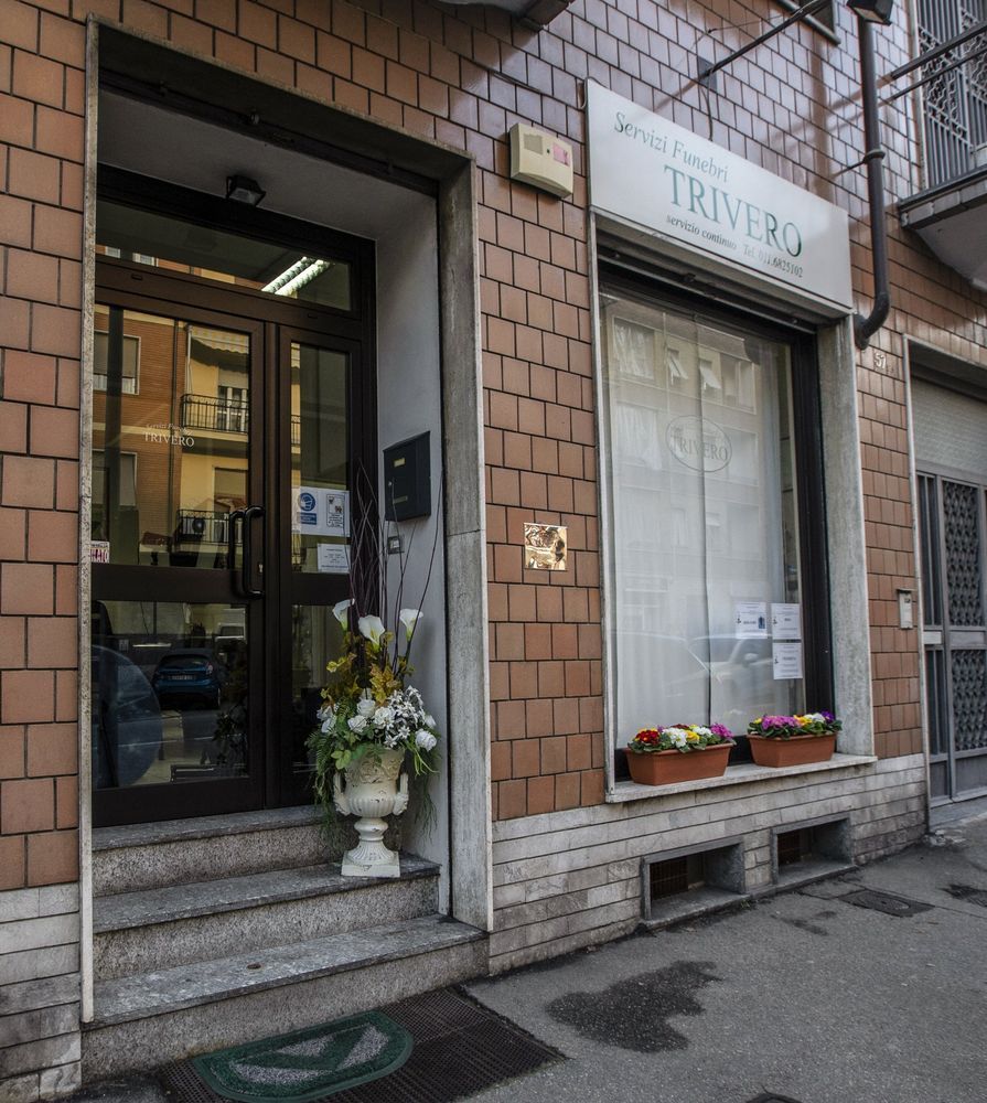 Negozio di piccole dimensioni con porta a vetri, facciata in mattoni, insegna TRIVERO e vasi di fiori vicino alla finestra.