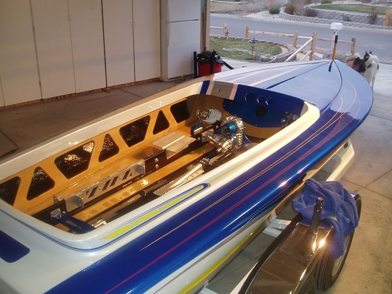 Blue-and-white racing powerboat on a trailer in a workshop, with wooden interior and docked water beyond