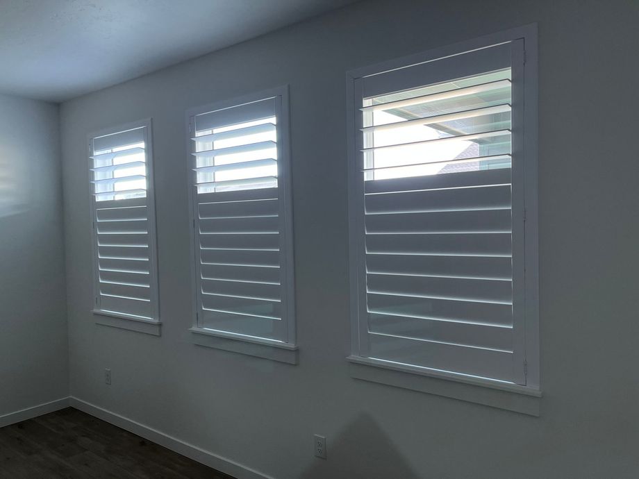 Three windows with white plantation shutters installed on a plain, light-colored wall.