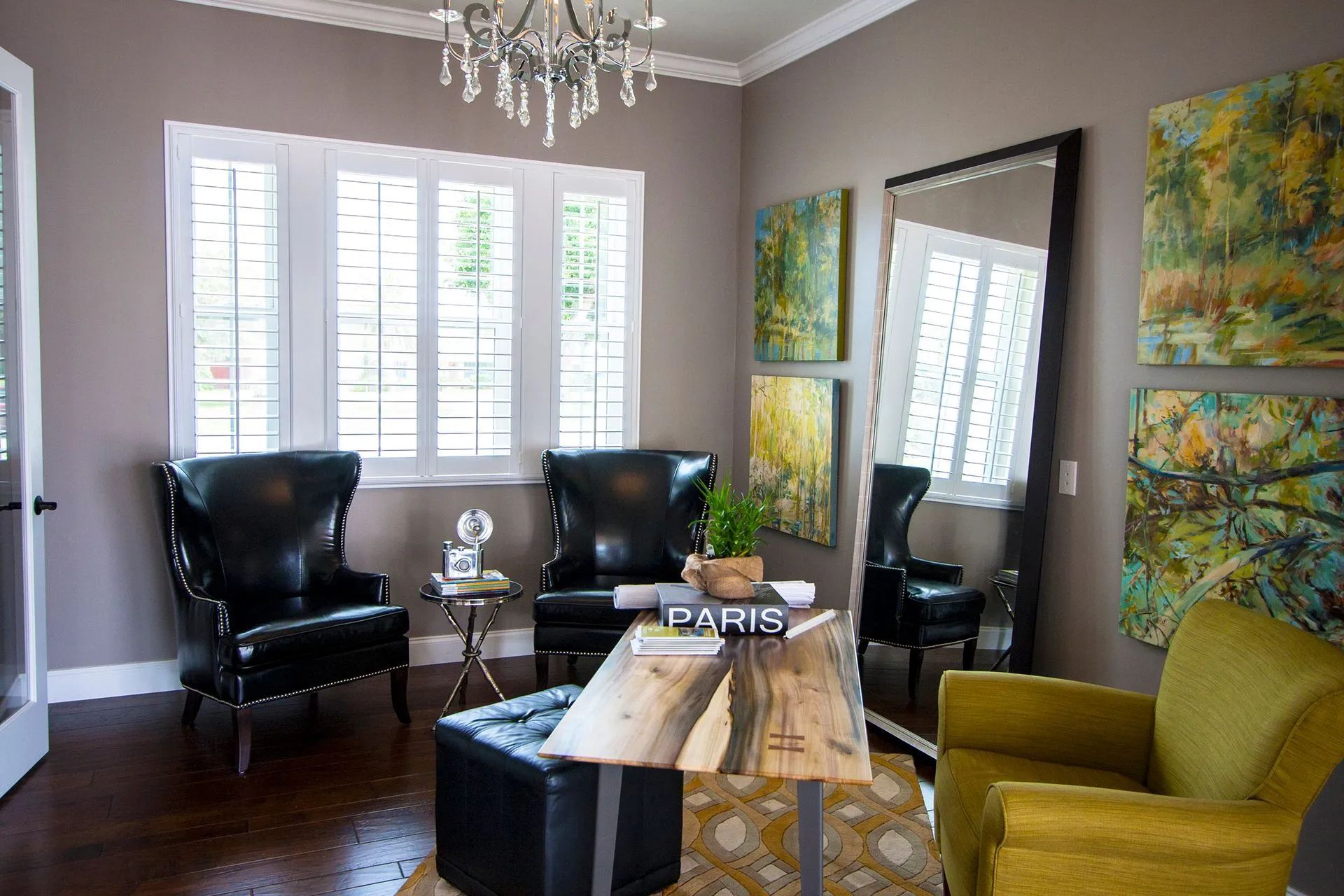 A home office with two black leather armchairs, a wooden desk, a black ottoman, and a large floor mirror with wall art.