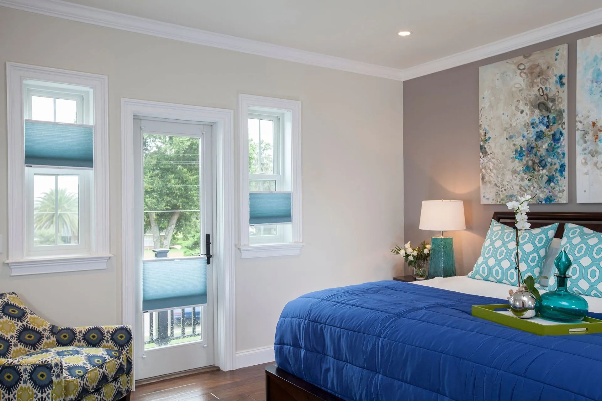 A bright bedroom with a blue bed, a floral armchair, and windows overlooking greenery.
