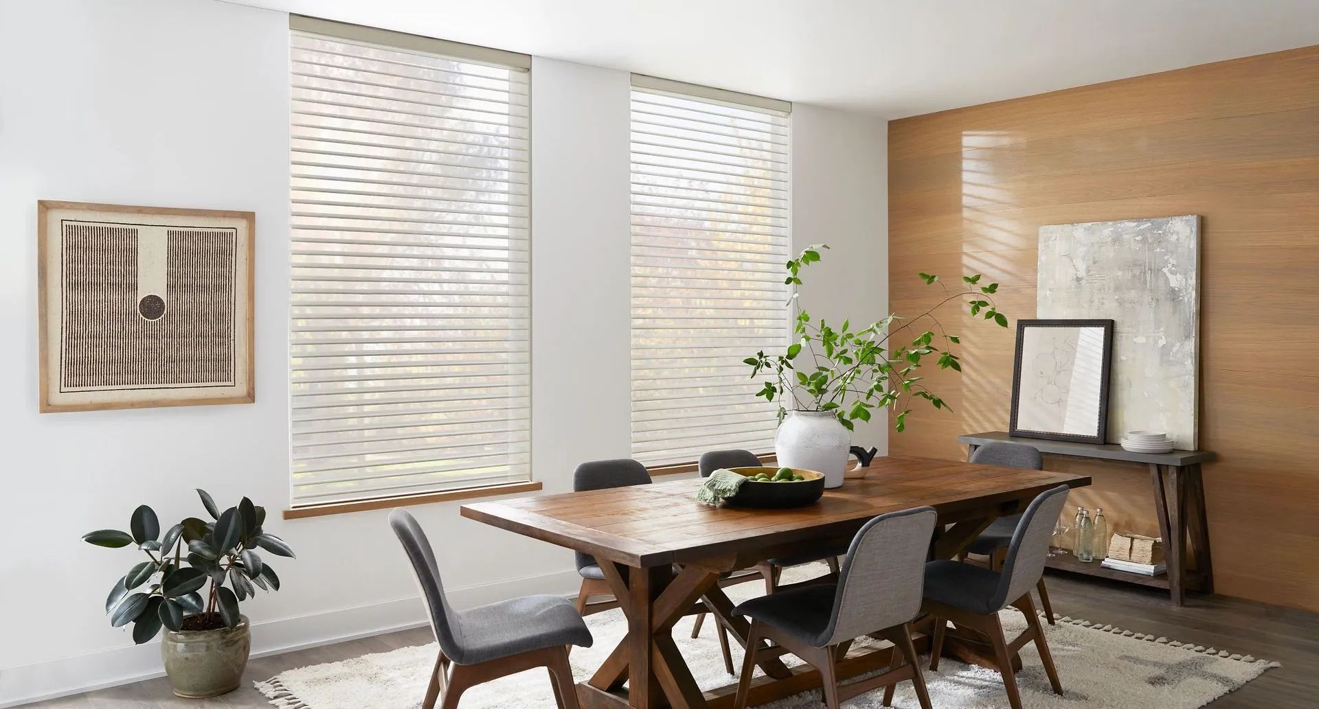 Dining room with a wooden table, grey chairs, indoor plants, and large windows with blinds against a wood-paneled wall.