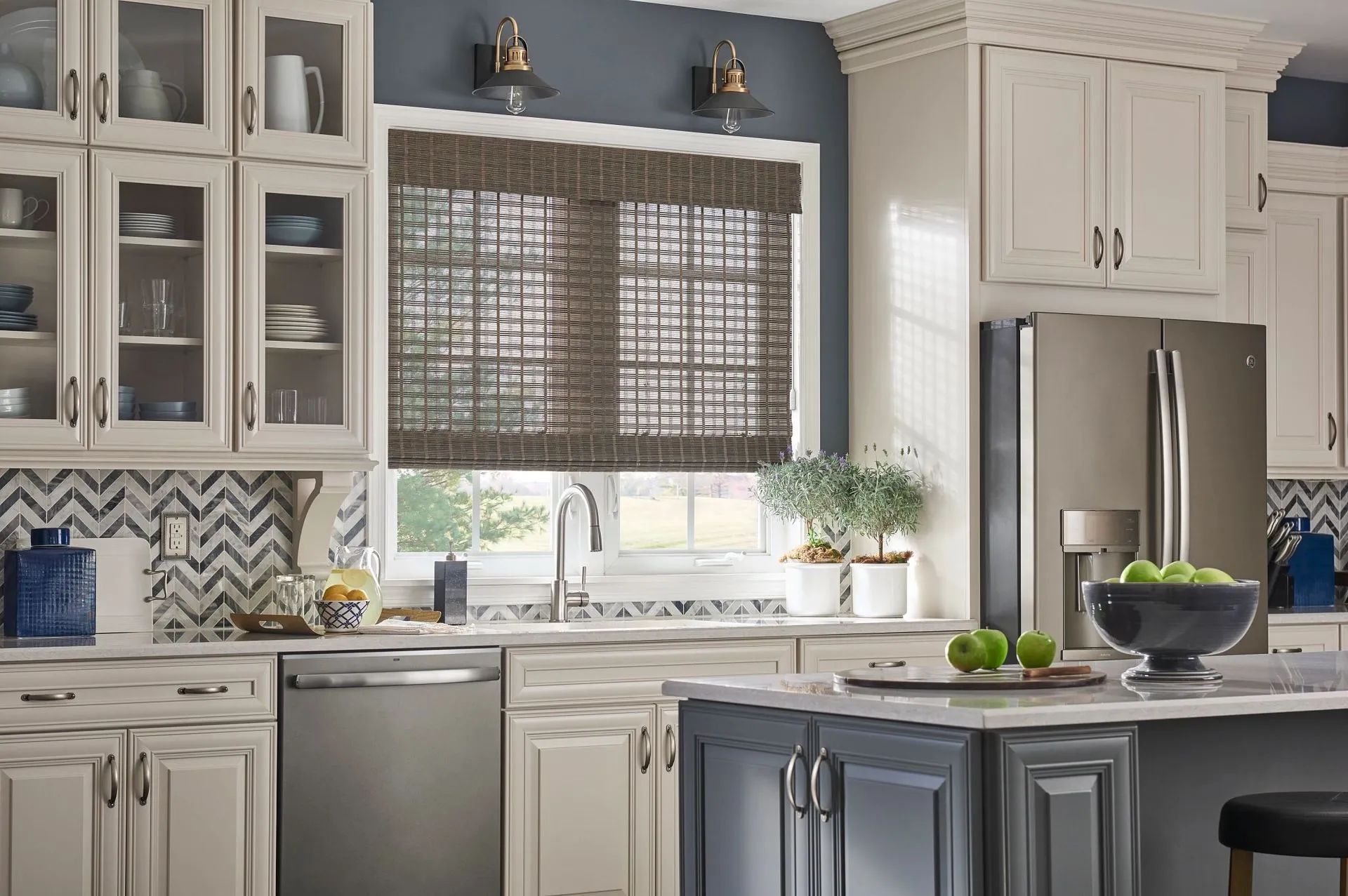 A modern kitchen with cream-colored cabinetry, a blue-gray island, stainless appliances, and woven wood window shades.