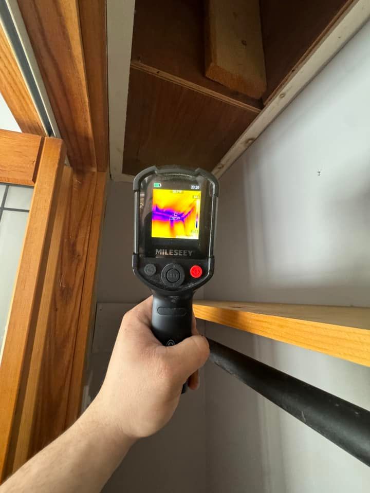 Handheld thermal camera aimed at a ceiling corner and wall, showing heat patterns on the screen