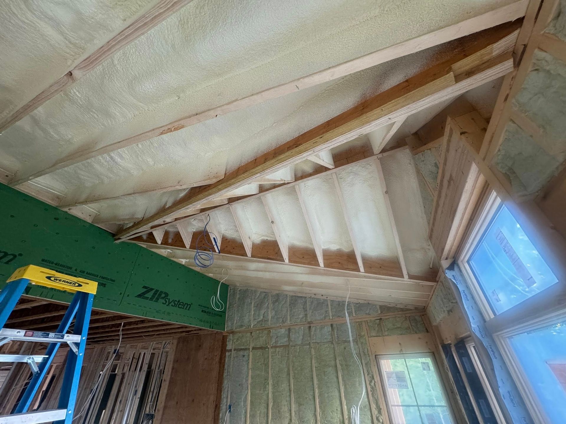 Partially insulated attic interior with exposed rafters, a blue ladder, and windows letting in daylight