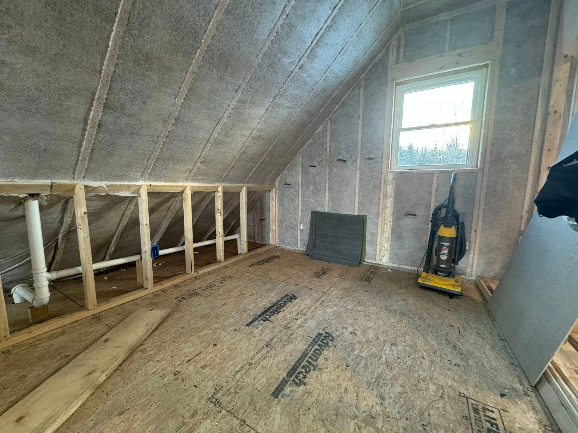Unfinished attic room with exposed framing, sloped ceiling, small window, and a yellow shop vacuum.
