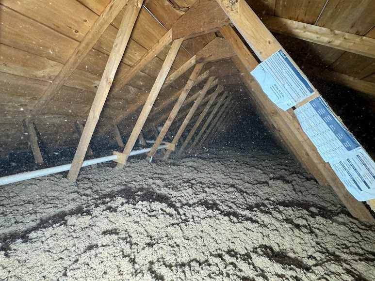 Inside an attic, exposed wooden rafters and a rough, dusty floor with insulation along the right side.
