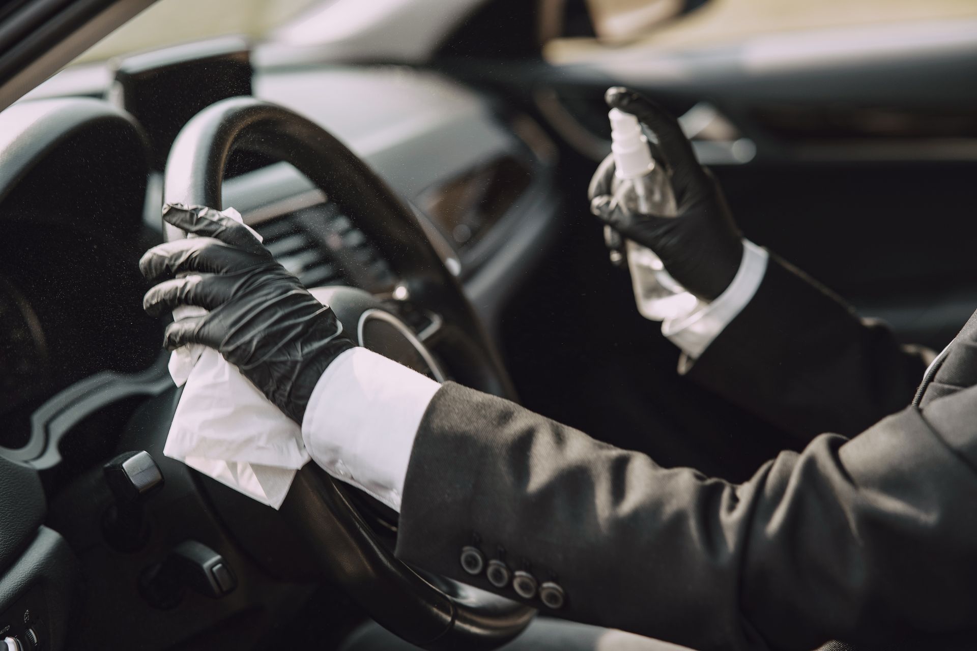Person wearing black gloves sprays and wipes the steering wheel of a car.