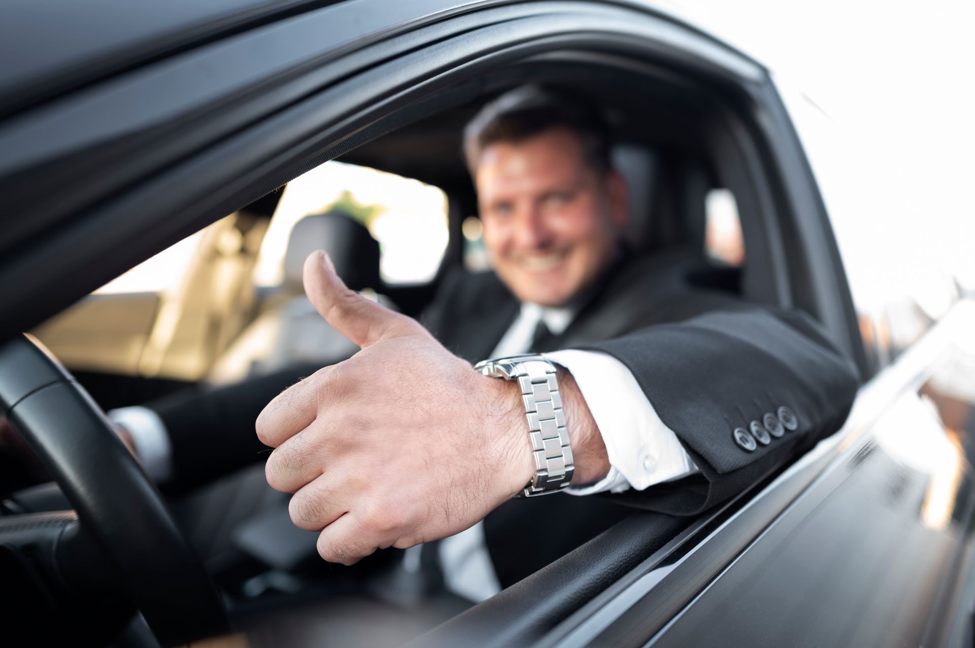 Man in a car giving a thumbs-up. He wears a suit and a watch, smiling.