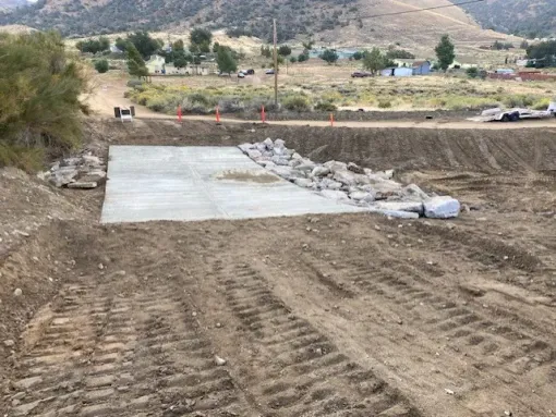 Construction site with a concrete pad and rocks. Dirt surrounding, hillside and buildings in the background.