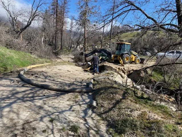 A person and a backhoe work on a dirt road in a wooded area, with a large hose visible.