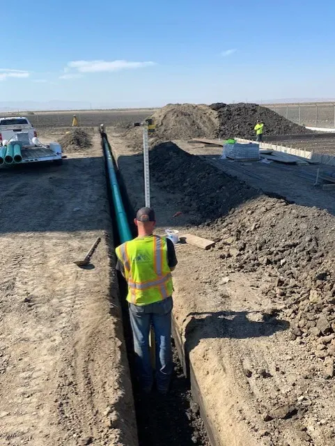 Construction worker in trench, installing pipe. Blue sky, open field, dirt, and equipment visible.