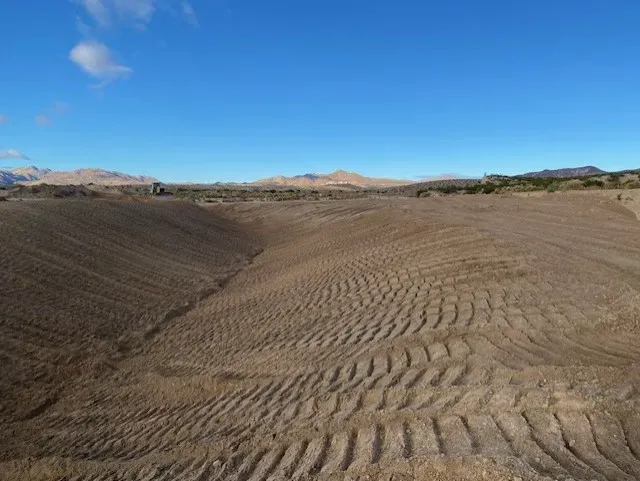 Brown, dry field with tire tracks and a drainage channel under a blue sky, mountains in the distance.