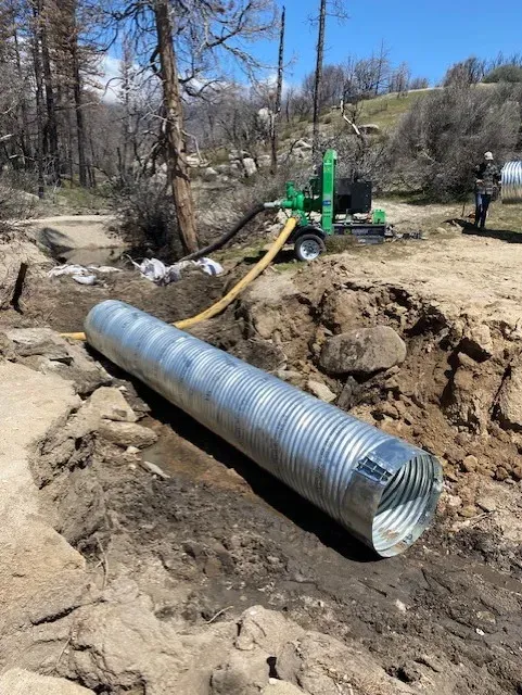 A corrugated metal culvert pipe lies in a trench with a pump in the background on a gravel road.