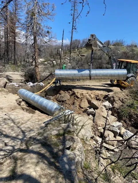 Excavator placing large corrugated metal pipe over a rocky creek bed, trees and blue sky background.