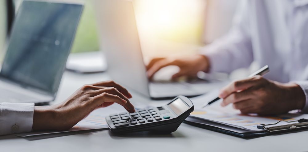 Hands using a calculator and writing on a clipboard, and using a laptop. Office setting.