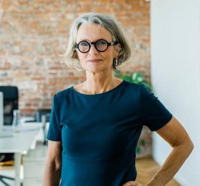 En kvinde iført briller og en blå skjorte står på et kontor med hænderne på hofterne.