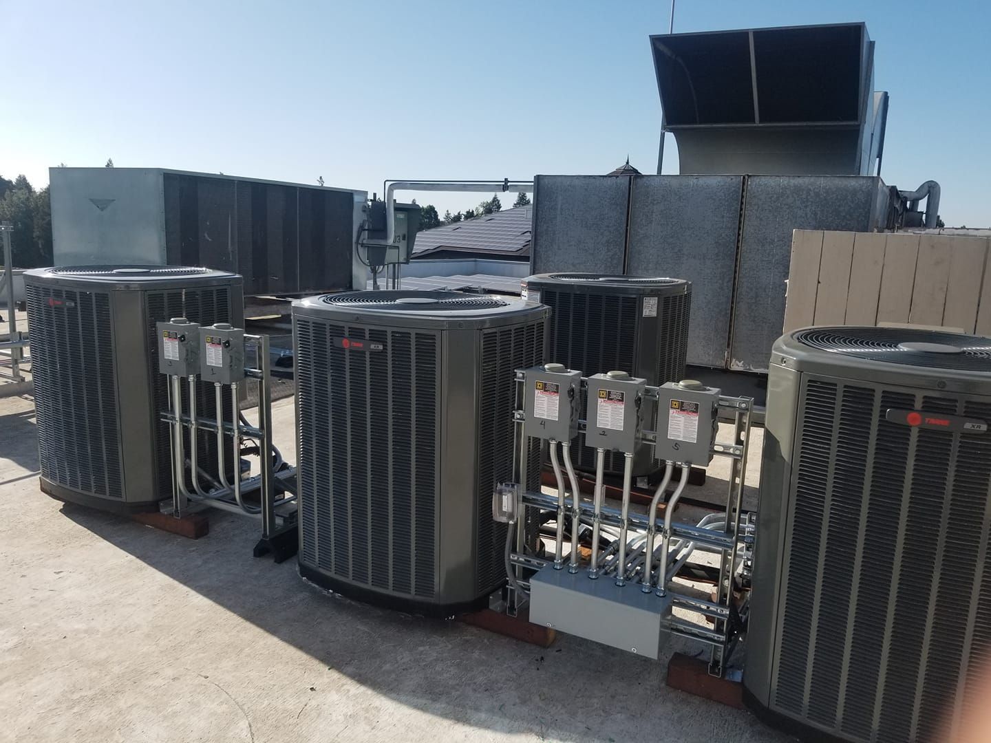 Several HVAC units on a rooftop, with electrical boxes and ductwork visible, under a clear sky.