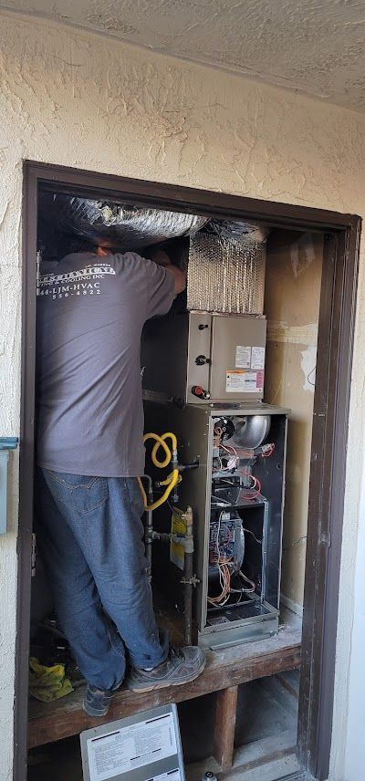 An HVAC technician works on a furnace inside a framed opening. He's wearing a gray shirt and blue pants.