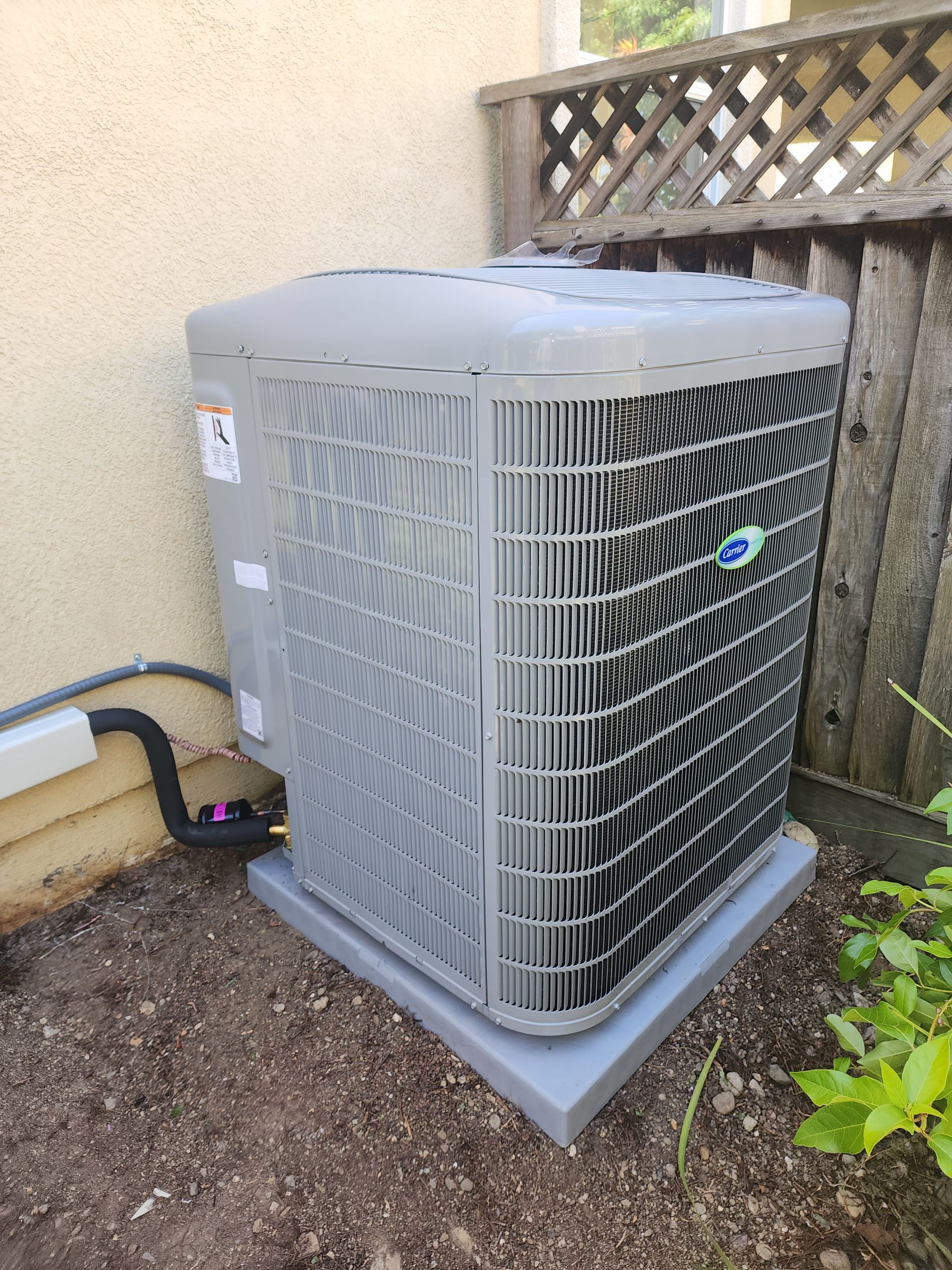 Gray air conditioning unit on a concrete pad next to a wall and a wooden fence.