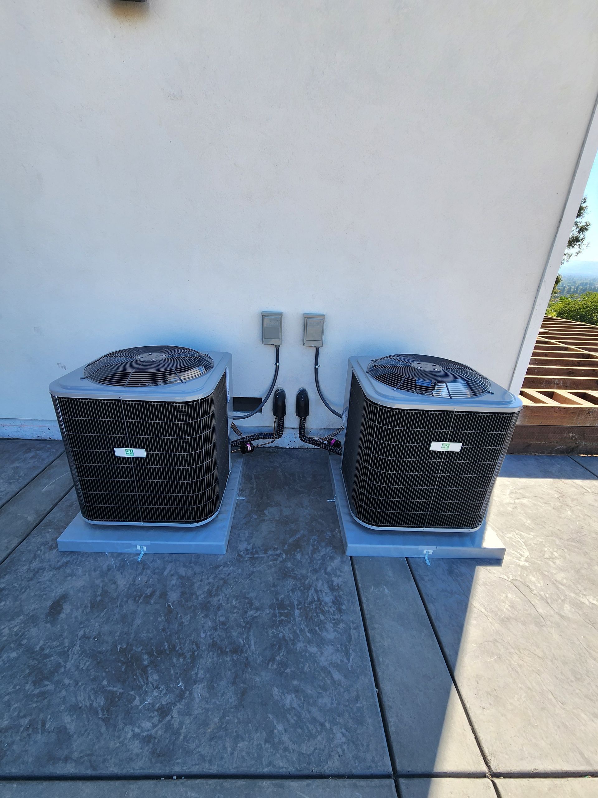 Two air conditioning units on concrete pads against a white wall.