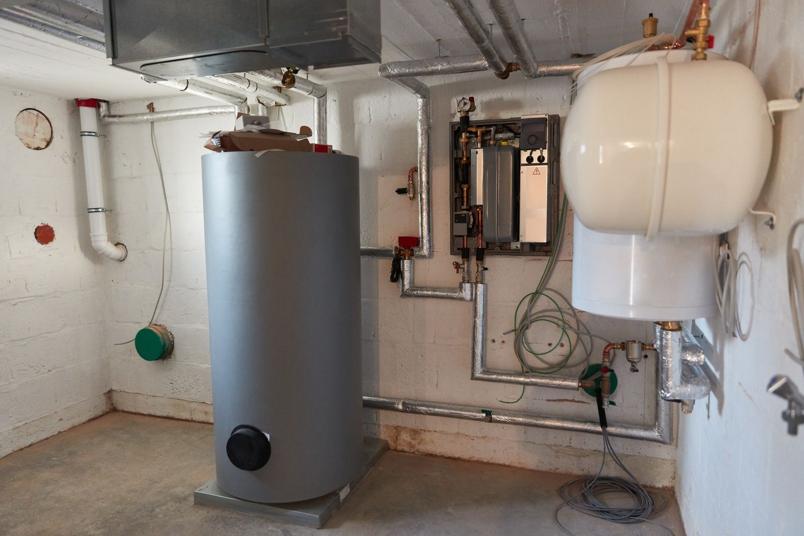 A utility room with a large water heater, expansion tank, and piping. White walls and gray floor.