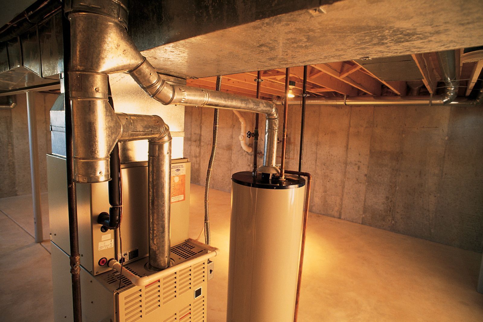 Furnace and water heater in a basement with silver ductwork against concrete walls.