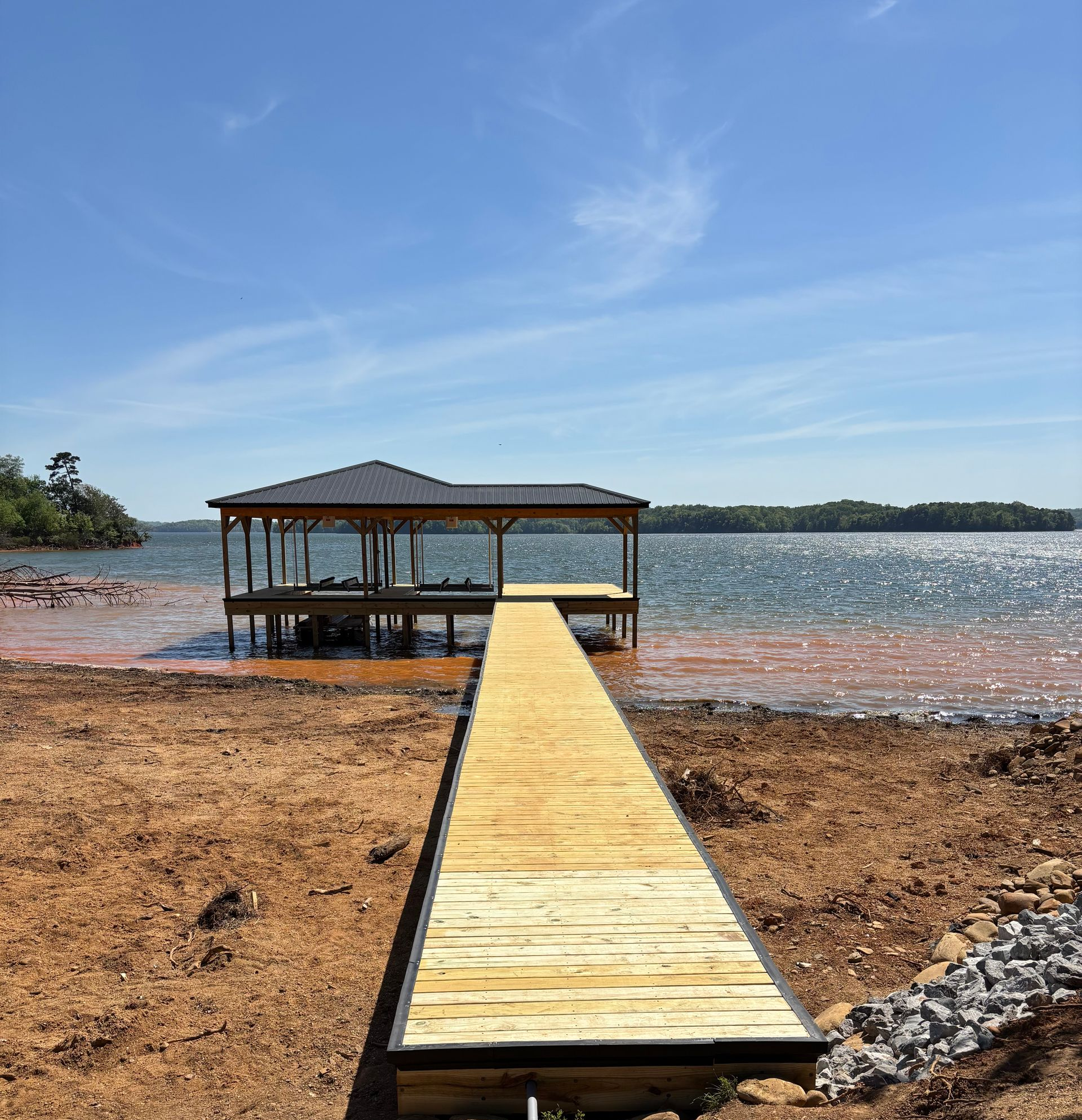 Wooden dock leading to a covered dock on a sunny shoreline, with blue water and sky.