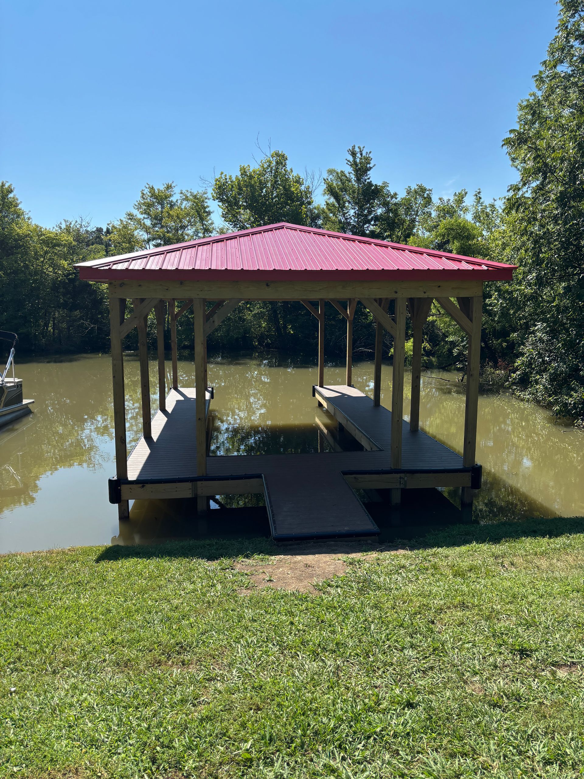 Dock with red roof, wooden beams, and boat slips in a waterway, surrounded by trees and green grass.
