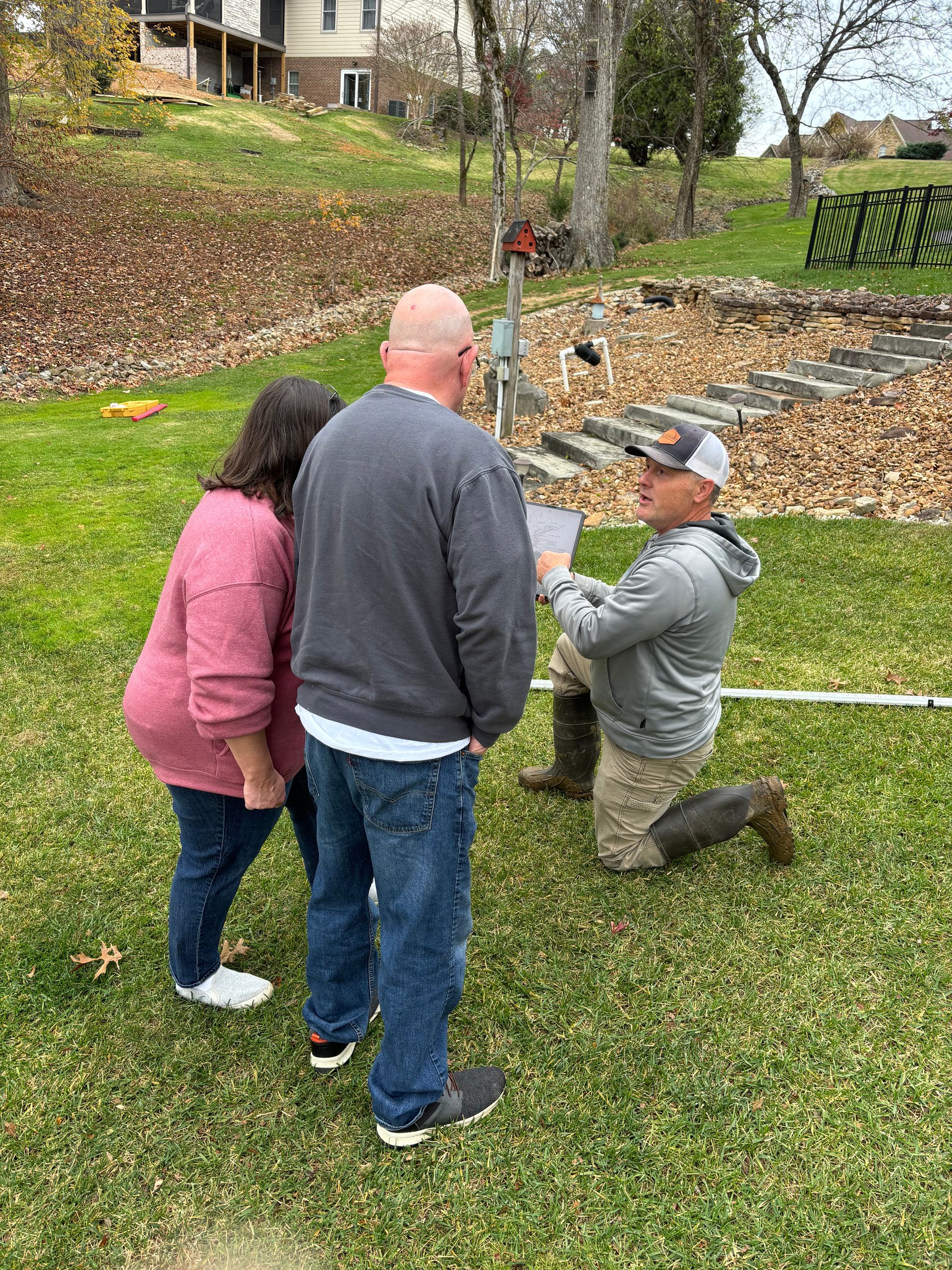 A man is kneeling down in the grass while talking to a couple.