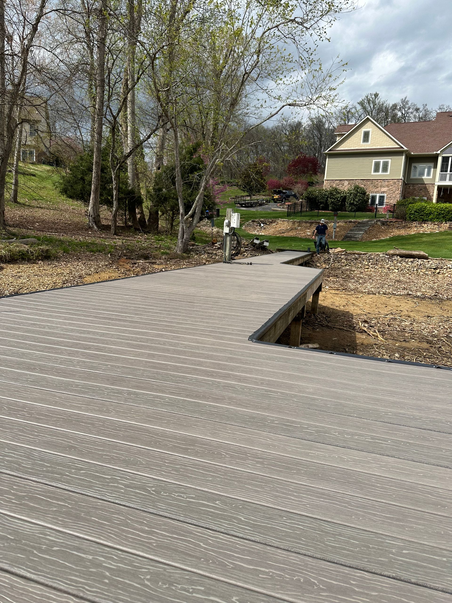 Gray composite dock extending into a lake, with trees and a house in the background.