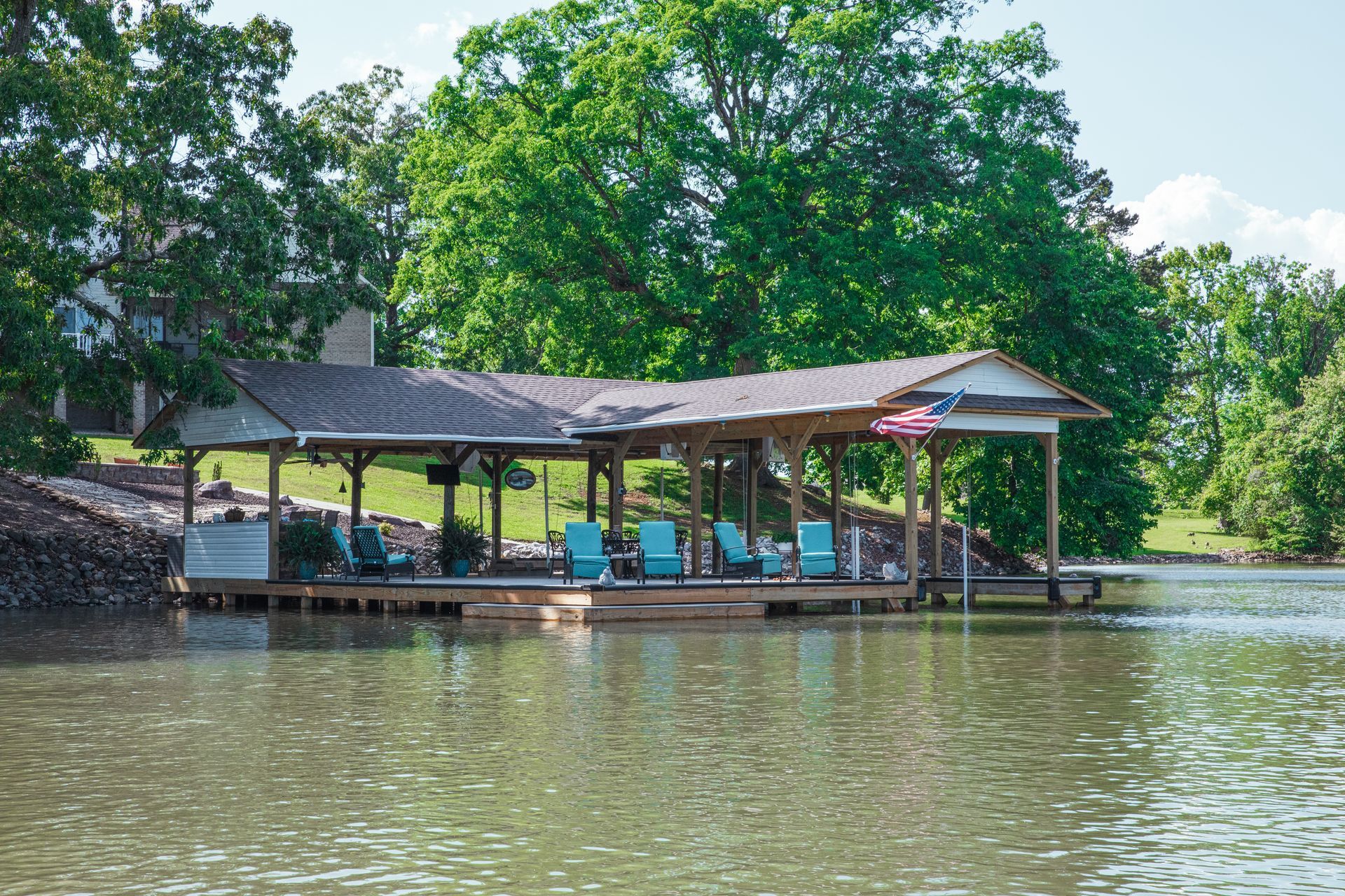 Lakeside pavilion with blue chairs on stilts over calm water, surrounded by green trees