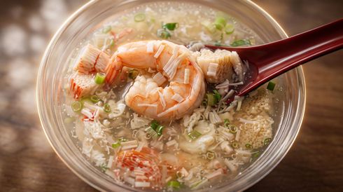 Shrimp congee in a clear bowl, topped with chopped scallions and a red spoon