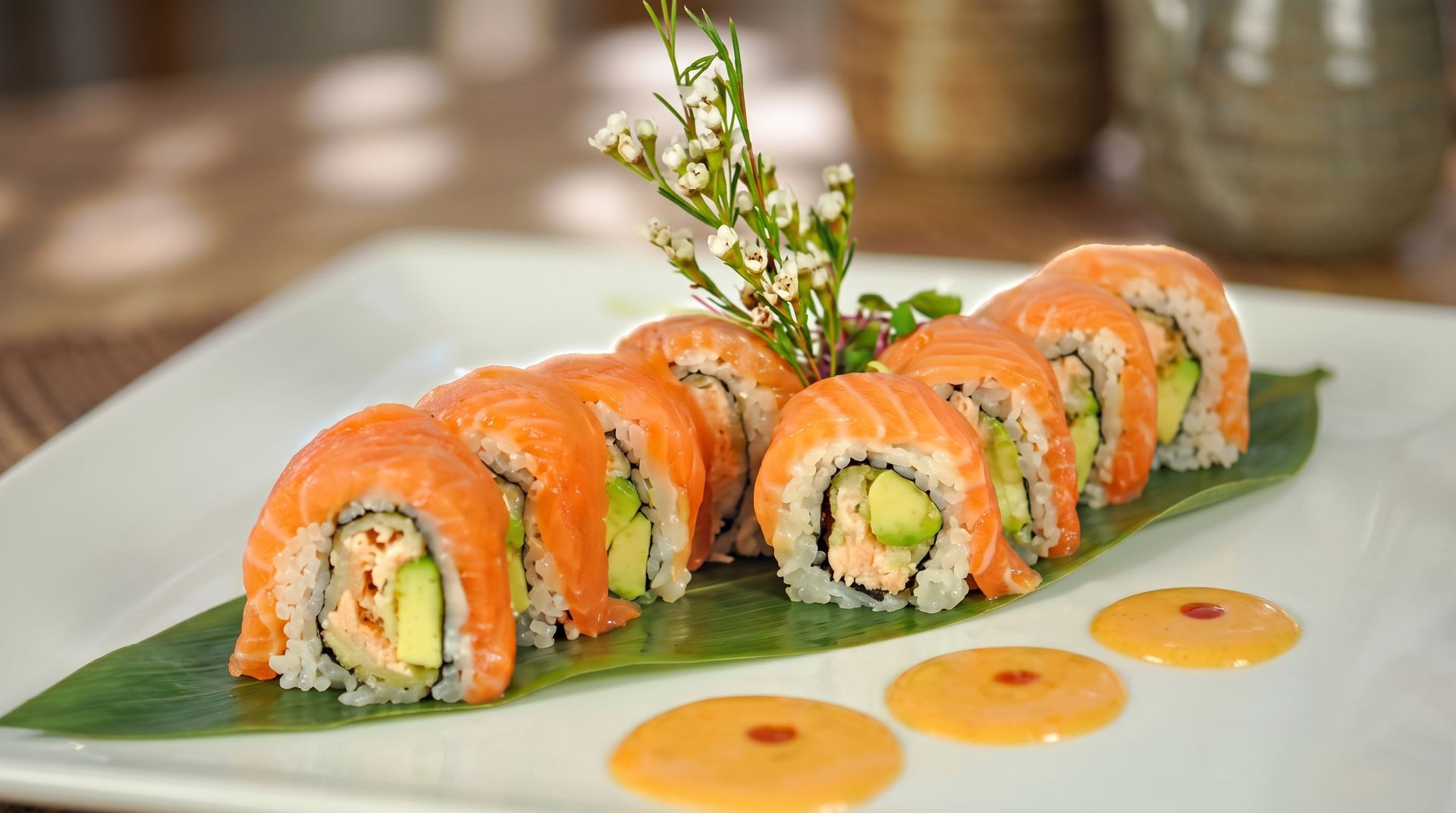 Sushi rolls topped with orange salmon on a white plate, garnished with herbs and sauce dots