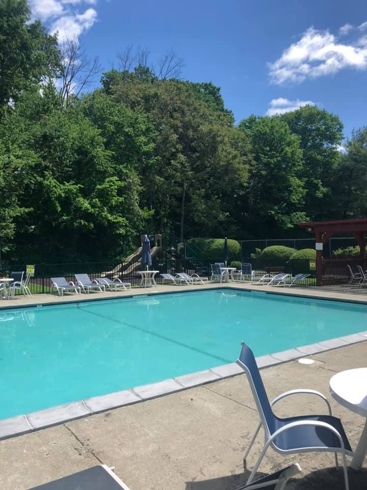 A rectangular outdoor swimming pool surrounded by lounge chairs, patio furniture, and lush green trees under a blue sky.