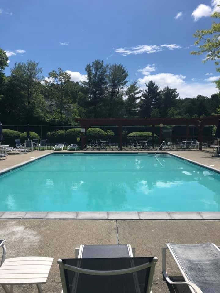 A bright turquoise swimming pool under a blue sky, viewed from a patio with lounge chairs in the foreground.