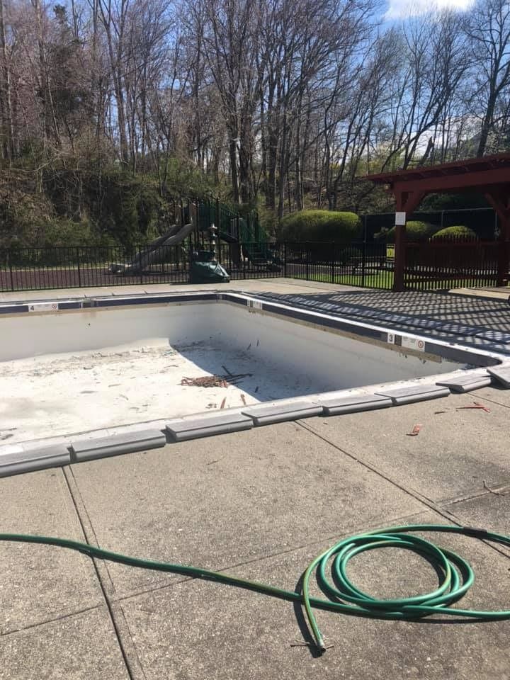 An empty, rectangular swimming pool surrounded by a concrete deck with a green garden hose lying in the foreground.